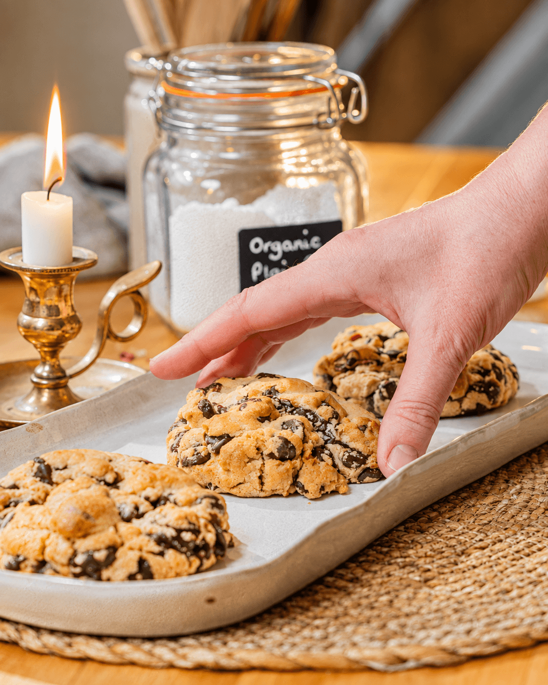 A hand reaching for a chocolate chunk cookie on a tray beside a candle and glass jar.