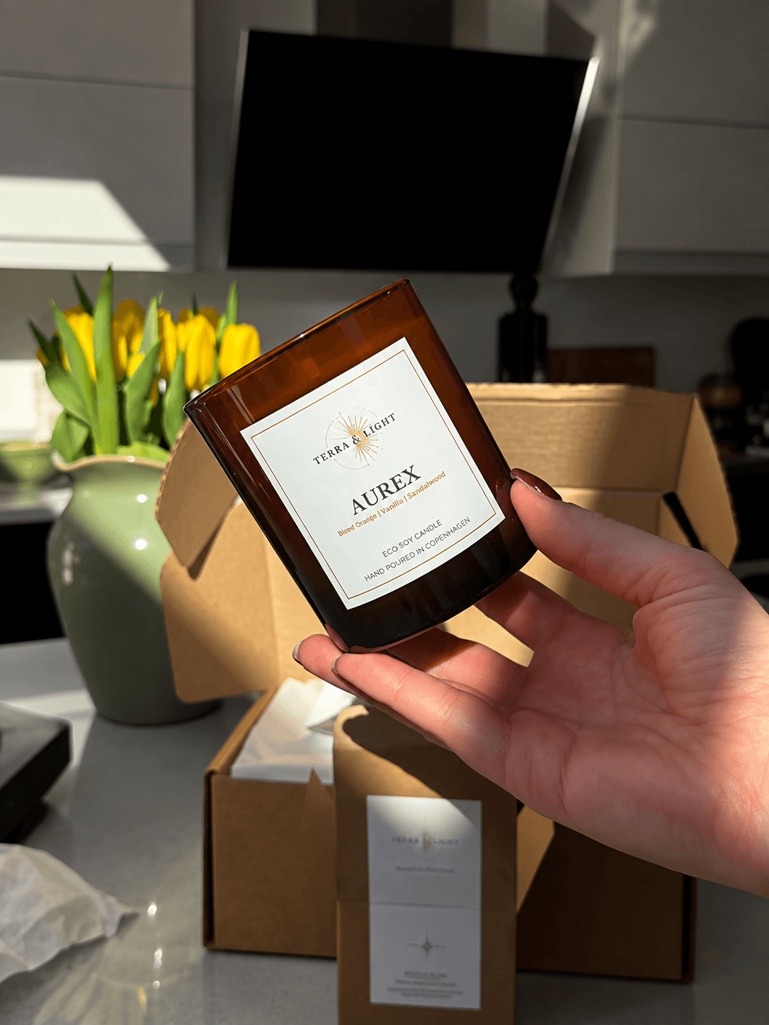 Hand holding an amber Aurex candle by Terra & Light beside an open gift box and yellow flowers.